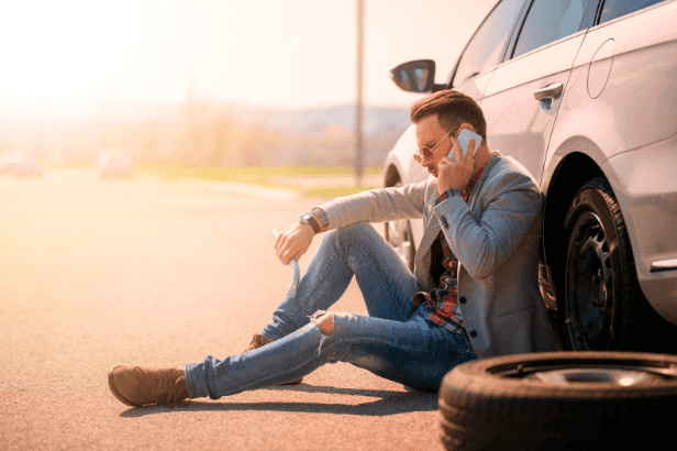 Roadside Assistance - Man in jeans sitting against his car that has a flat tire