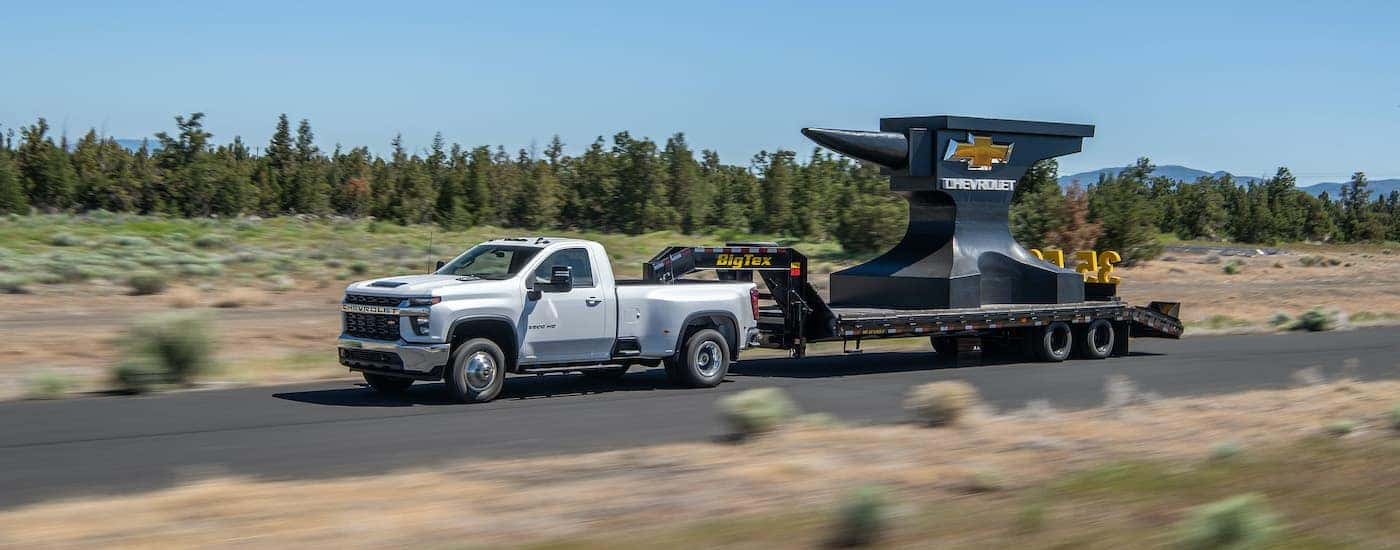 A white 2022 Chevy Silverado 3500HD is shown towing a giant anvil on an open road.