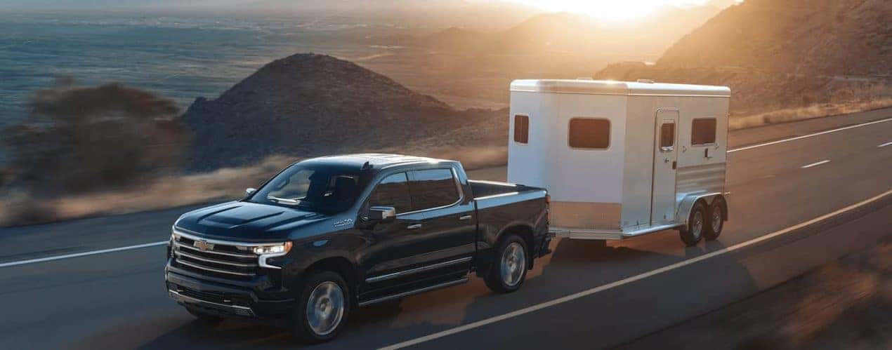 A grey 2023 Chevy Silverado 1500 High Country is shown towing a white trailer on a highway.