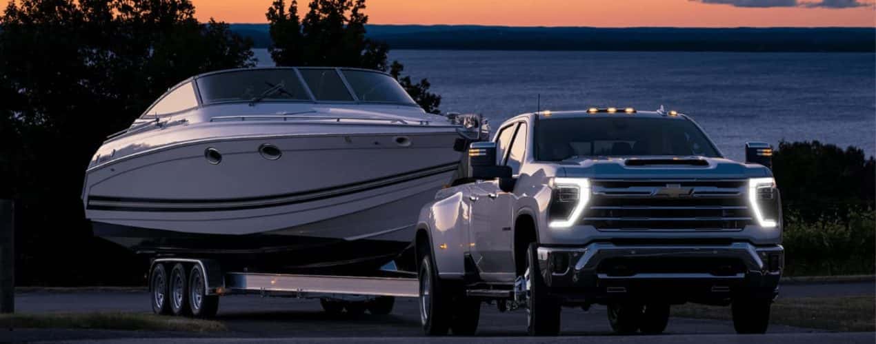 A white 2024 Chevy Silverado 3500 HD is shown towing a boat during a sunset.