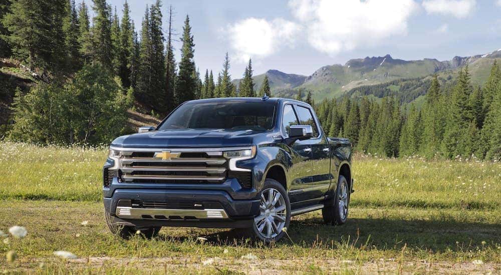 A blue 2022 Chevy Silverado 1500 High Country parked in a grassy field with trees and mountains