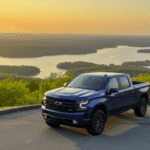 A magnificent 2026 Chevrolet Silverado pickup truck, gleaming in a deep metallic finish, stands proudly at golden hour, parked on a paved overlook high above the expansive, calm waters of Lake Guntersville, Alabama, its powerful silhouette dominating the foreground, showcasing its modern design and rugged capability. The scene is bathed in the warm, soft glow of a spring sunset, with an orange and purple sky reflecting gently on the lake's surface, surrounded by vibrant, lush green trees and emerging foliage indicative of early April, capturing the serene beauty of the southern landscape. The composition is eye-level, emphasizing the truck's impressive stature against the breathtaking scenic backdrop, evoking a sense of adventure, power, and aspirational lifestyle, with rich colors and exquisite detail in a photorealistic style.