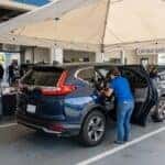 A realistic photograph of a family SUV parked under a canopy tent in a dealership service drive during a community safety clinic, a child safety seat being secured in the second row, orange cones and folding tables nearby, sunny late-morning light, wide 16:9 composition. No text, no logos other than natural vehicle badging, no identifiable faces. (Representative image for illustration purposes only. Actual vehicle may vary based on trim and configuration.)