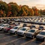 A realistic photograph of rows of new pickup trucks and SUVs lined up on a dealership lot at golden hour, autumn hardwood trees along the back fence, long shadows across the pavement, wide 16:9 composition. No text, no logos other than natural vehicle badging, no identifiable faces. (Representative image for illustration purposes only. Actual vehicle may vary based on trim and configuration.)
