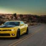 Yellow and black 2015 Chevrolet Camaro driving down a paved road past tall mountains and plants