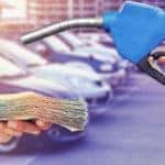 Hand holding blue gas nozzle toward another hand with cash in it, in front of parked cars