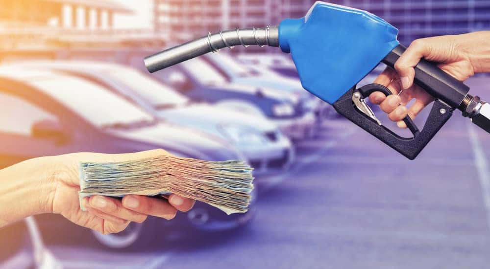 Hands exchanging cash and a gas nozzle in front of a parking lot filled with cars