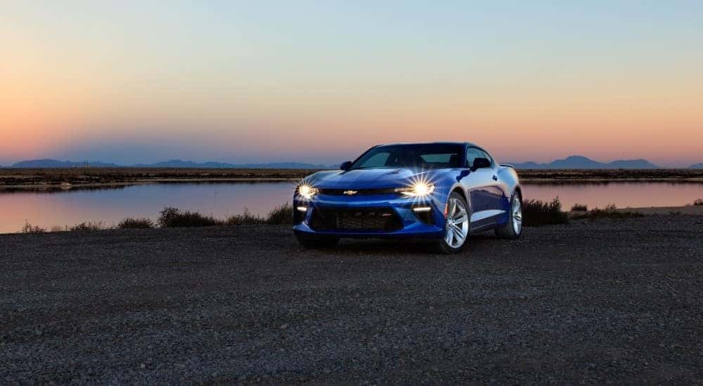 Front view of a blue 2016 Chevrolet Camaro sports car parked in front of a body of water and a sunset