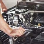 Hands with tools leaning against the front of a car with its hood open