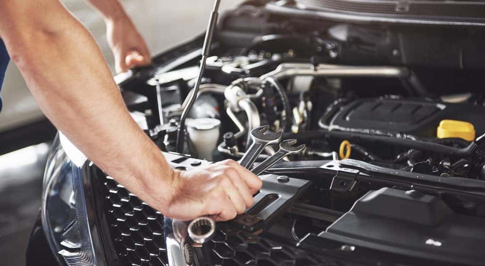 Hands with tools leaning against the front of a car with its hood open