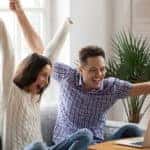 Man and woman sitting on a couch with their arms up in excitement in front of a brown coffee table with a laptop on it