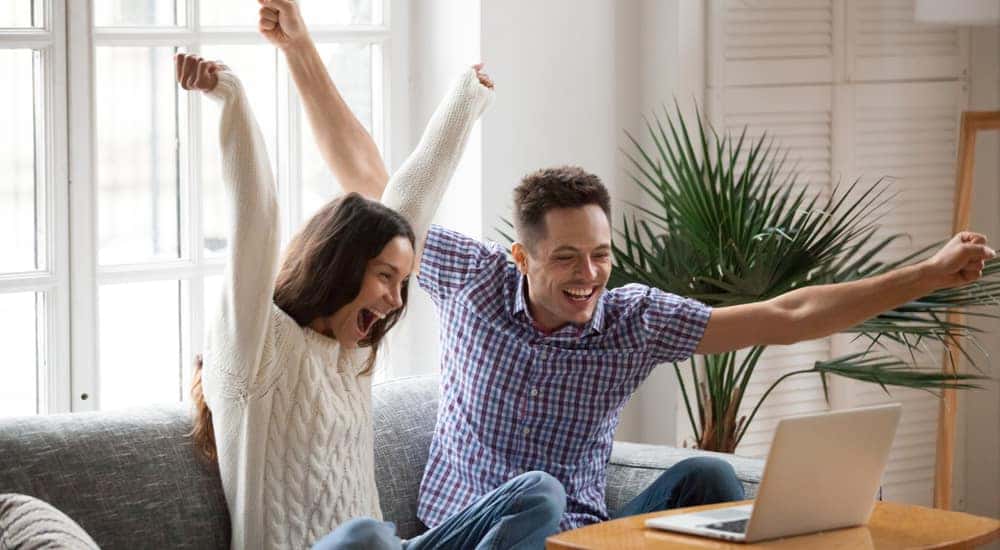 Man and woman sitting on a couch with their arms up in excitement in front of a brown coffee table with a laptop on it