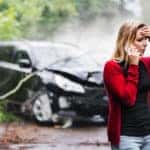 Woman in a red cardigan distressed on the phone in front of her smashed up vehicle