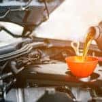 Hands pouring oil from a grey bottle into the tank under a vehicle's hood through an orange funnel