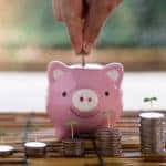 Stacks of coins with green plants coming out of the top sitting in front of a pink piggybank on a brown surface