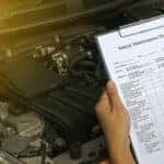 Hands holding a clipboard with a vehicle maintenance checklist on it above the open hood of a vehicle