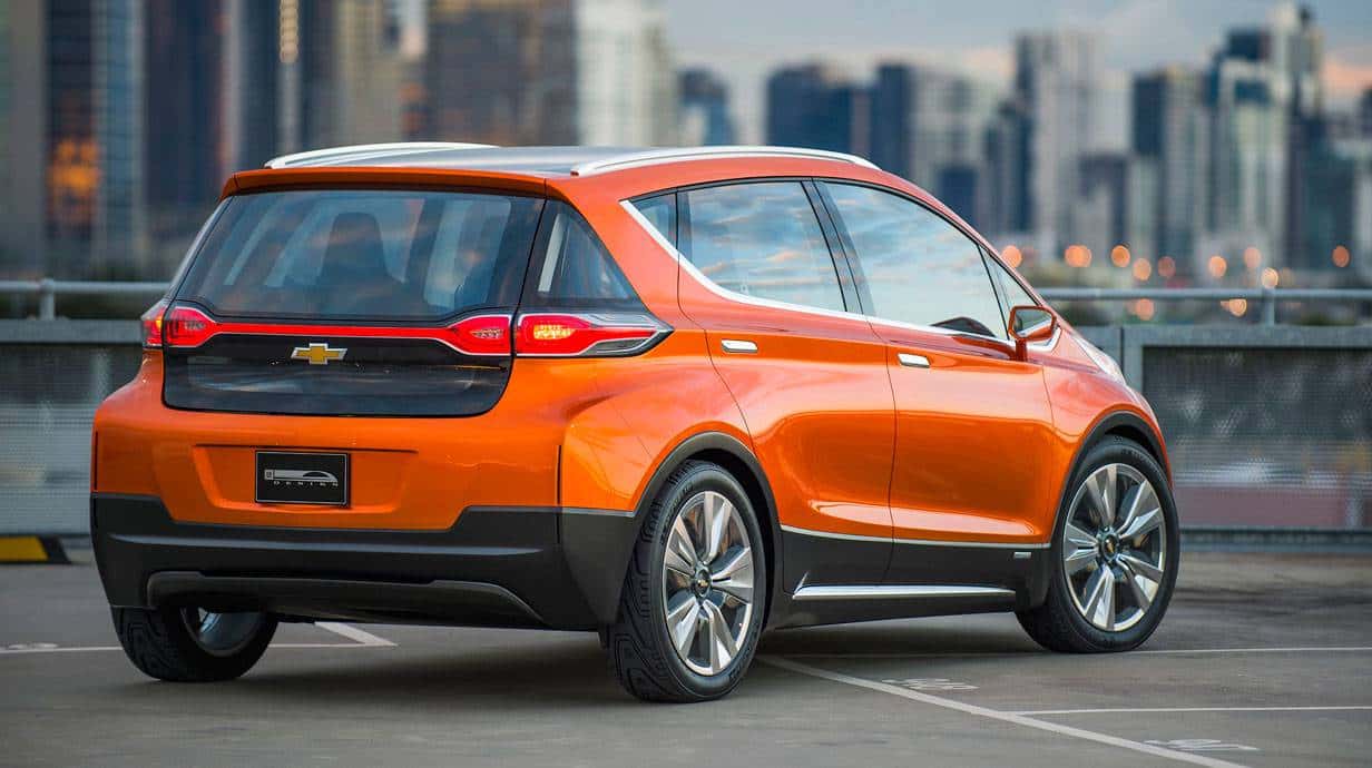An orange 2015 Chevy Bolt is shown in a a parking lot with a city in the distance.