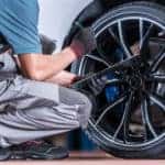 Man in a light blue shirt and grey overalls bending down and inspecting tires on a silver vehicle