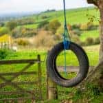 Tire hanging from a tree with a blue rope in front of a wooden gate and tall green hills