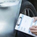 Man writing on a clipboard and assessing a large dent in a silver vehicle