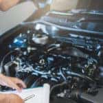 Mechanic looking under the hood of a vehicle with a clipboard in hand inside an auto repair shop