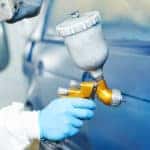Person in a white suit and mask painting a blue vehicle with a professional paint tool
