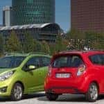 One green and one red Chevrolet Spark hatchback parked in front of water and a city skyline