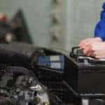 Male mechanic in a blue jumpsuit changing the battery under the hood of a car