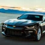 Black 2016 Chevrolet Camaro sports car driving on a paved road next to a body of water and blue mountains