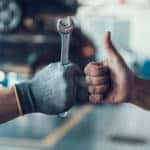 Hand giving a thumbs up next to a hand in a glove holding a tool in front of cars in a mechanic's shop