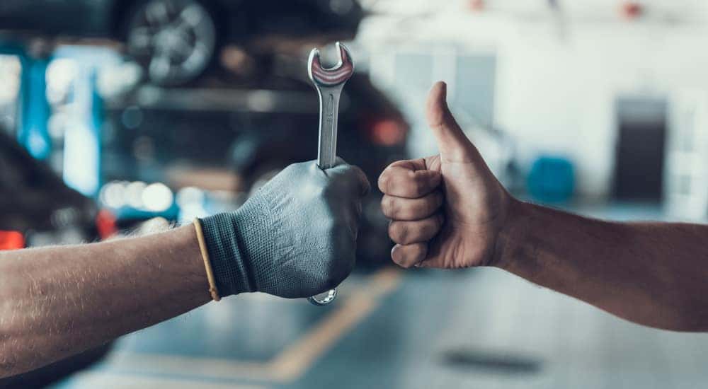 Hand giving a thumbs up next to a hand in a glove holding a tool in front of cars in a mechanic's shop