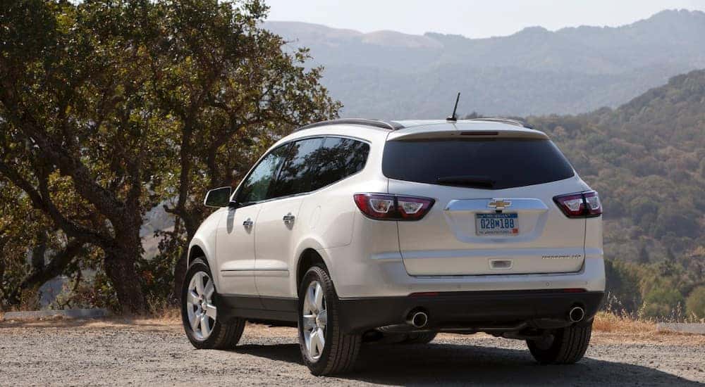 Back to front view of a white 2016 Chevrolet Traverse SUV parked on a dirt ground in front of tall mountains