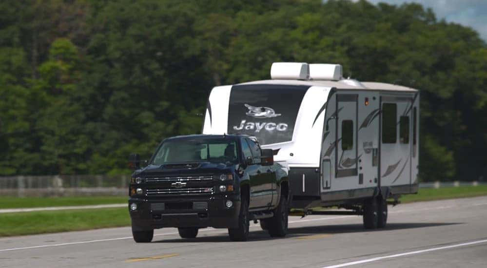 Black Chevrolet Silverado 2500HD truck, with a white camper attached, driving down a paved road
