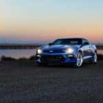 Blue 2016 Chevrolet Camaro with its headlights on parked in front of a body of water and sunset