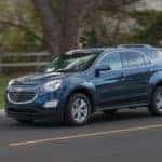Blue 2016 Chevrolet Equinox driving down a paved road past grass, a brown fence, and a tree