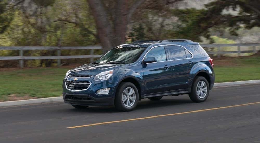 Blue 2016 Chevrolet Equinox driving down a paved road past grass, a brown fence, and a tree