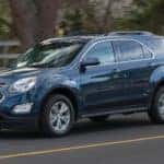 Blue Chevrolet Equinox driving on a paved road next to a tree, grass, and a white fence