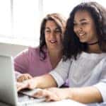 Teen with curly black hair sitting at a table with her mother and a white laptop