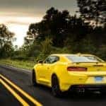 Yellow 2016 Chevrolet Camaro SS performance vehicle driving up a paved road past green grass and trees