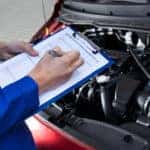 Man in a blue jumpsuit holding a blue clipboard with white paper on it standing over the open hood of a car