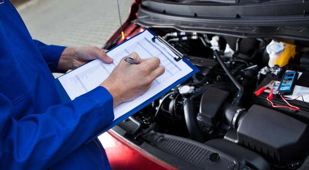 Man in a blue jumpsuit holding a blue clipboard with white paper on it standing over the open hood of a car