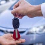 Hand of someone wearing a white button-up handing a car key to another person's hand in front of a sedan