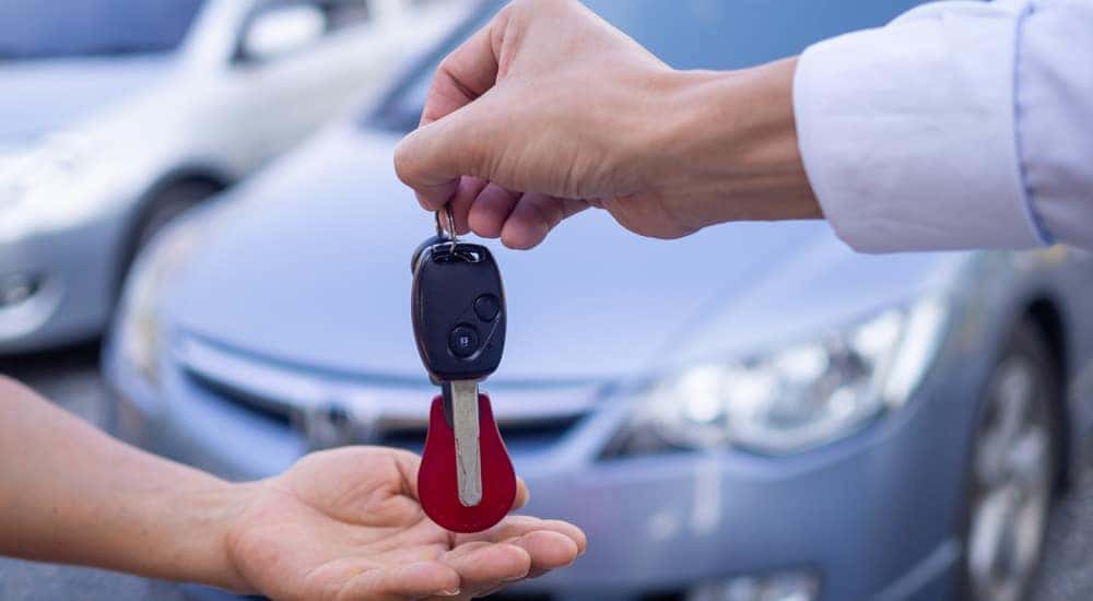 Hand of someone wearing a white button-up handing a car key to another person's hand in front of a sedan