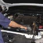 Man in a blue and grey short sleeve shirt working under the open hood of a white vehicle