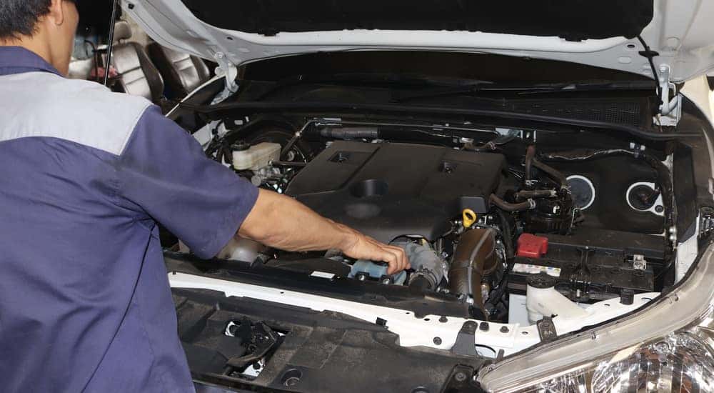 Man in a blue and grey short sleeve shirt working under the open hood of a white vehicle