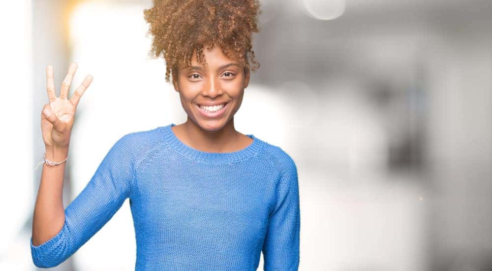 Woman in a blue sweater holding up three fingers in front of a blurred background