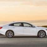 White 2016 Chevrolet Malibu parked on the side of a paved road in front of a large body of water