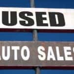 White sign that says "USED" on it and grey sign that says "Auto Sales" on it, both on a grey pole against a blue sky