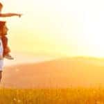 Mom and dad standing together with a daughter on the dad's shoulders in a field at sunset