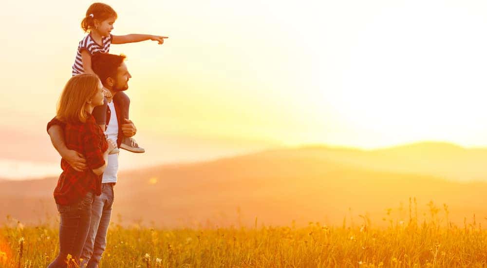 Mom and dad standing together with a daughter on the dad's shoulders in a field at sunset